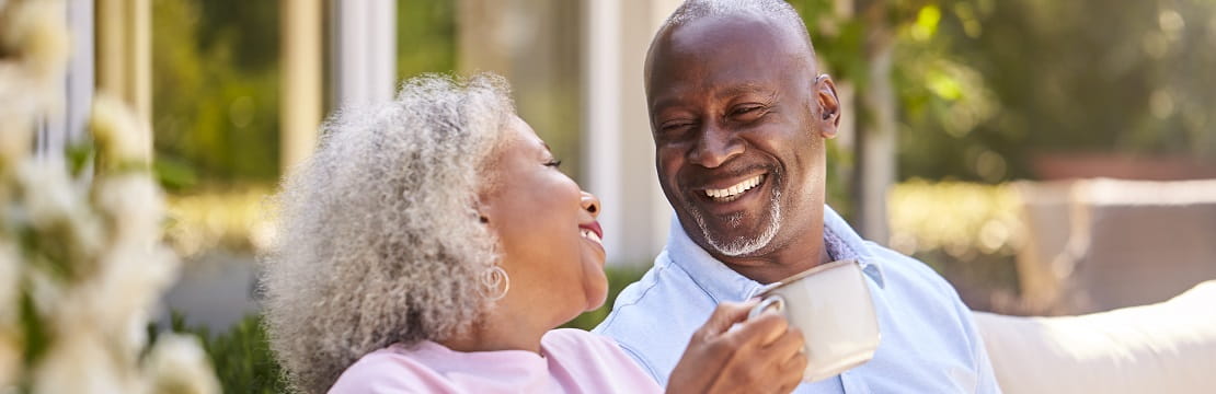 Senior couple smiles at each other while drinking out of coffee mugs. 